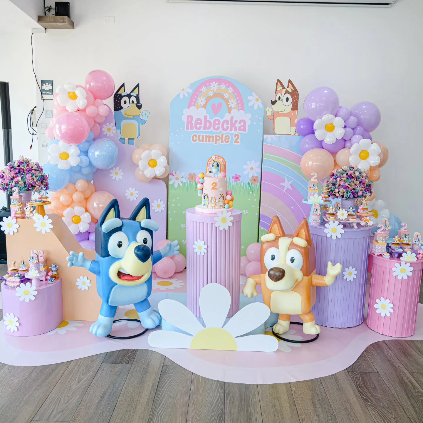 A full Bluey birthday party setup with a pastel rainbow backdrop, balloon garlands, and large Bluey and Bingo character figures.