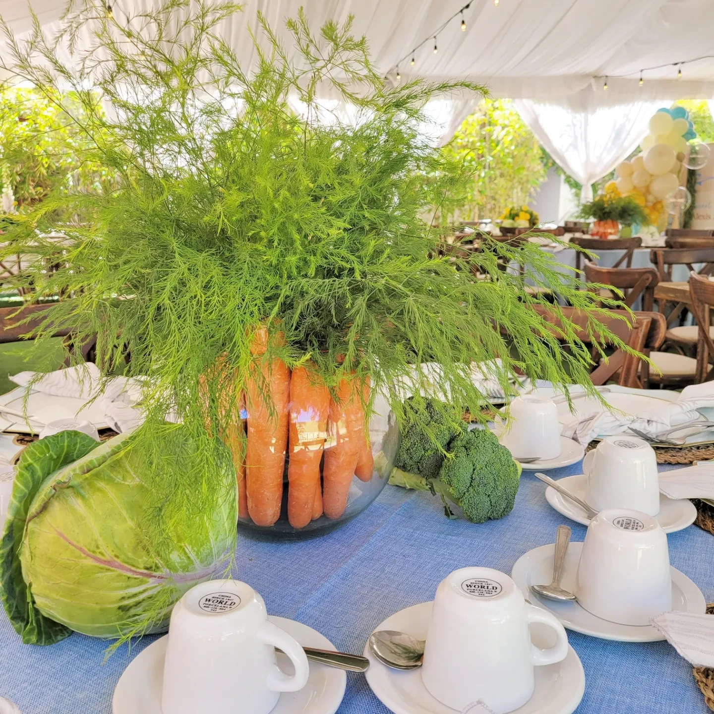 A Peter Rabbit baby shower centerpiece made from a glass bowl filled with carrots and topped with green ferns on a blue tablecloth.