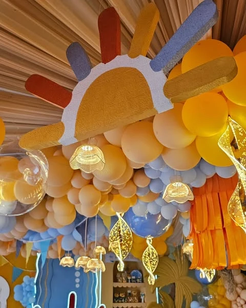 A of a large sun cutout with colorful rays hanging from the ceiling amidst yellow balloons for a baby shower.