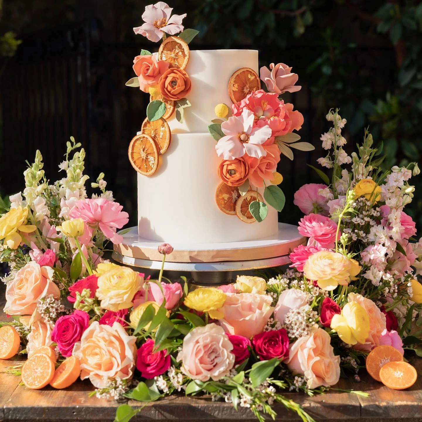 A two-tier white bridal shower cake decorated with dried orange slices and pink and orange sugar flowers, surrounded by an arrangement of fresh roses.