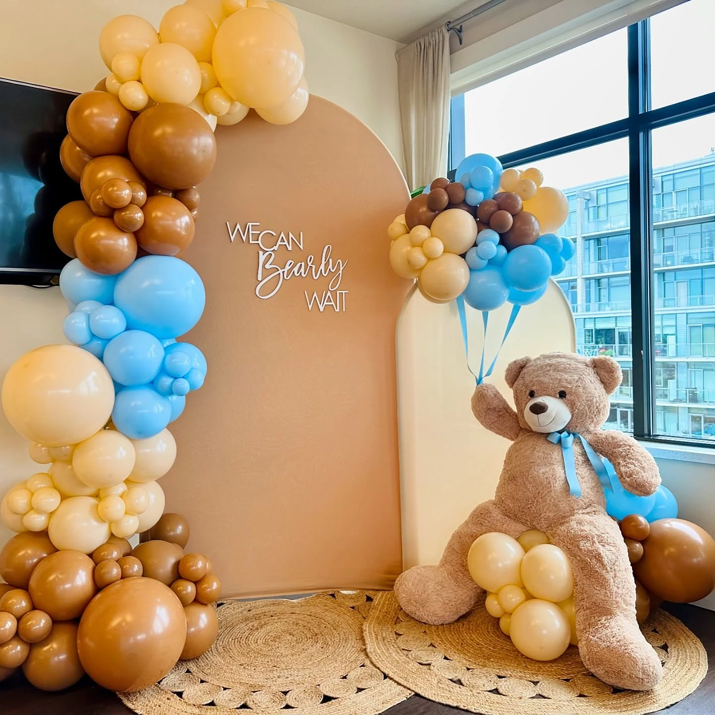 A 'We Can Bearly Wait' baby shower backdrop with a large teddy bear, arched panels, and a blue and brown balloon garland.