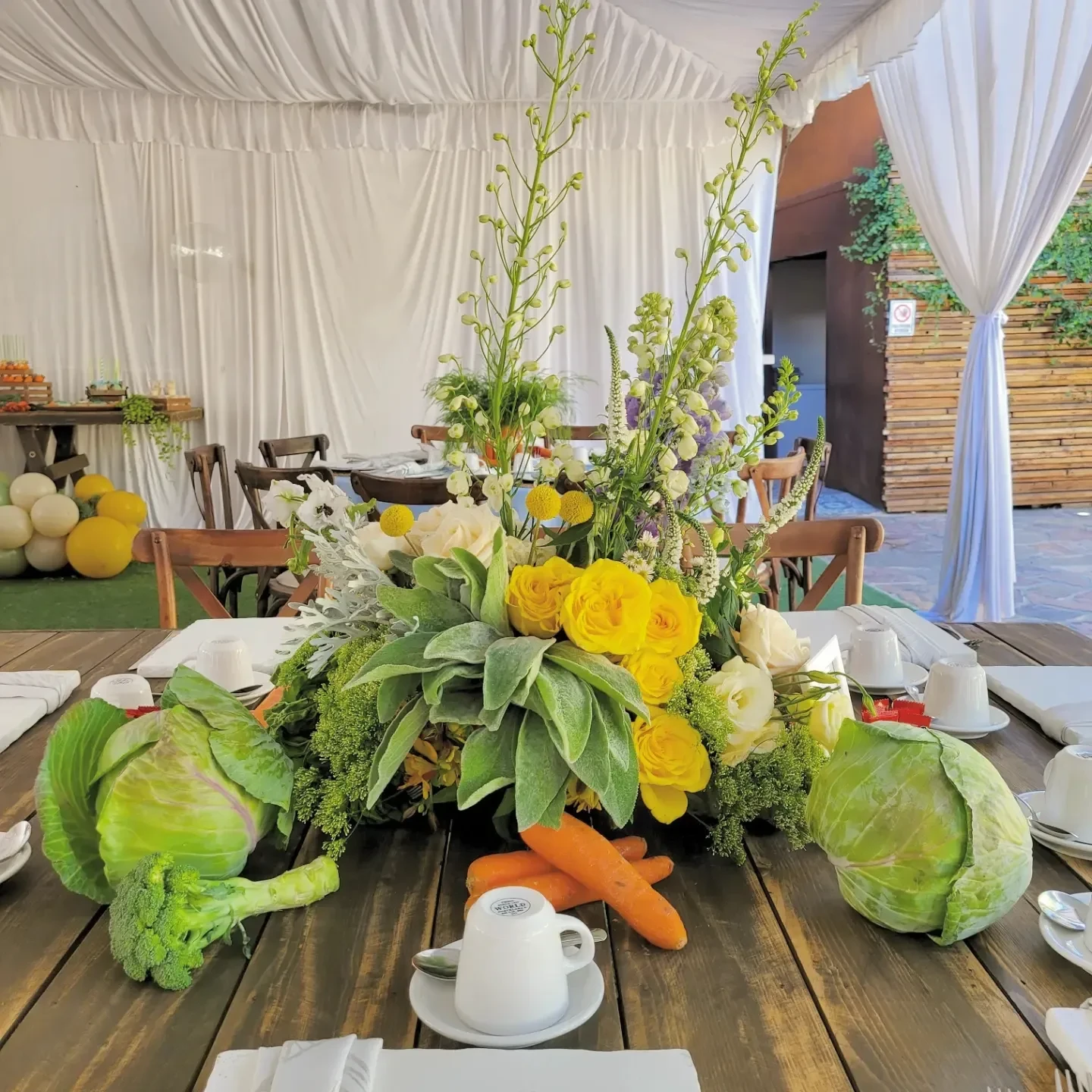 A floral centerpiece with yellow roses surrounded by fresh cabbages and carrots on a rustic wood table for a Peter Rabbit baby shower.