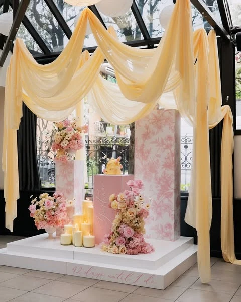 A romantic spring bridal shower backdrop featuring pale yellow draped fabric and pastel pink and cream floral arrangements.