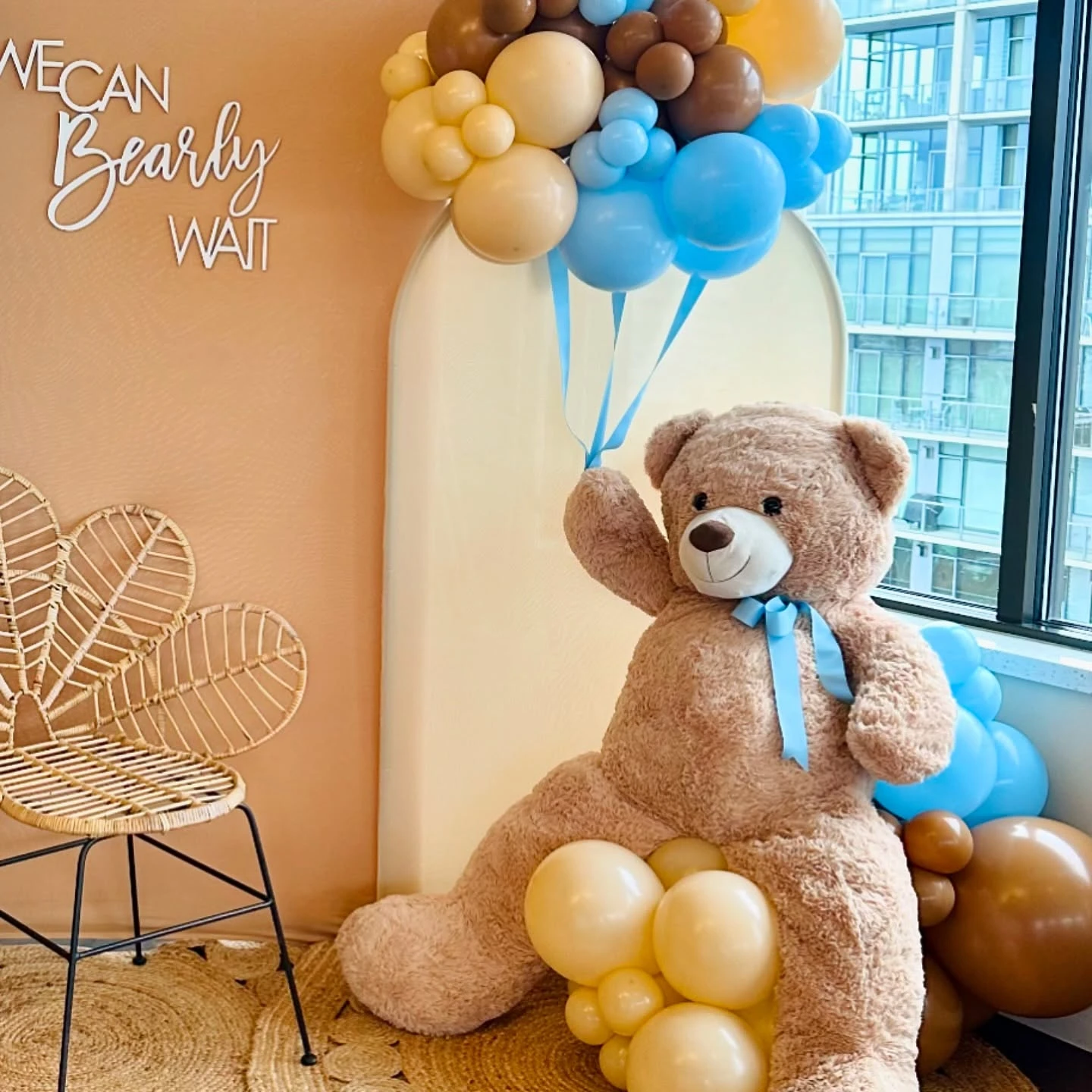 of a large teddy bear holding blue and brown balloons at a 'We Can Bearly Wait' baby shower.