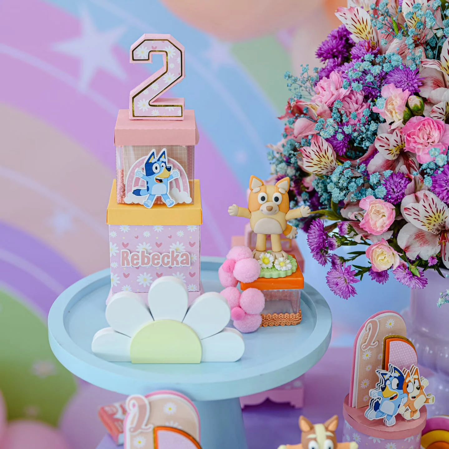 A Bluey party decor detail with a Bingo figure, a bouquet of flowers, and themed favor boxes on a light blue cake stand.