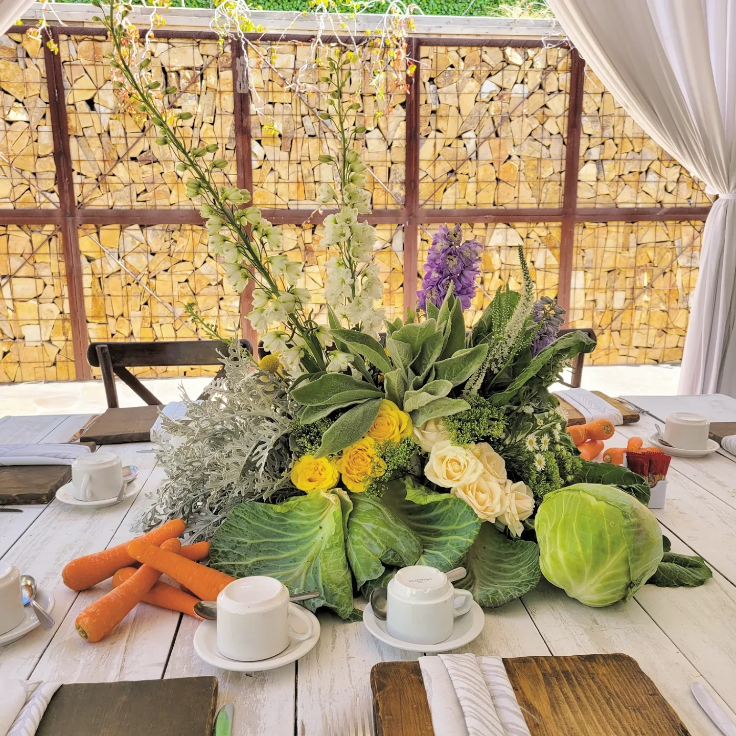 A garden-style centerpiece with flowers and vegetables, including large green cabbages, on a white wooden table.