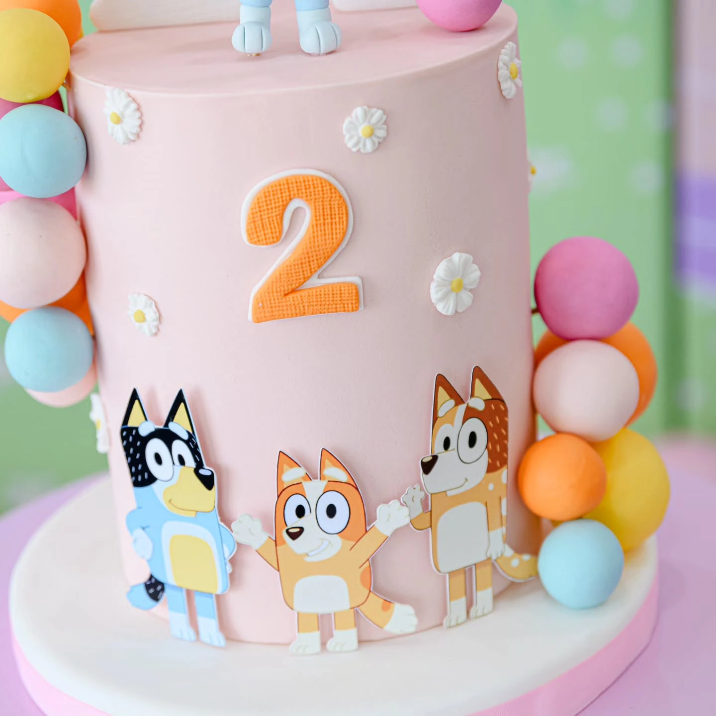 A pink Bluey-themed birthday cake with edible character cutouts and small fondant daisies.