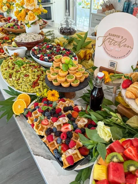 A of a baby shower food table featuring a platter of waffles with berries, mini quiches, and a pasta salad.