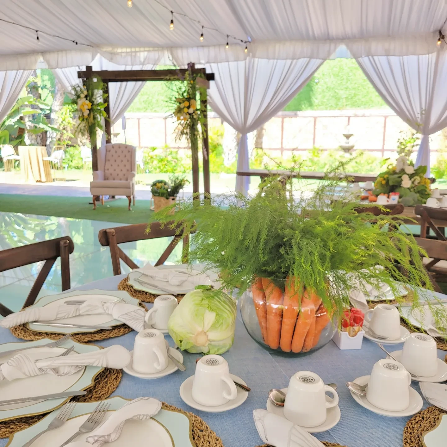 A full table setting for a Peter Rabbit baby shower featuring a centerpiece of carrots in a glass bowl on a blue tablecloth.
