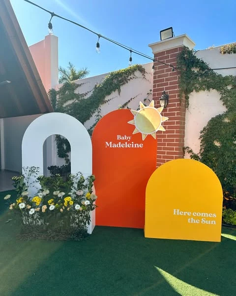 A modern 'Here comes the Sun' baby shower photo backdrop with orange, yellow, and white arched panels and a sun cutout.