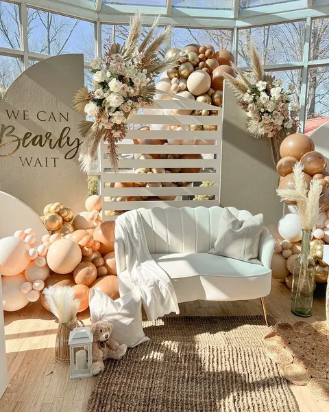 A 'We Can Bearly Wait' baby shower photo backdrop with a neutral balloon arch, pampas grass floral arrangements, and a small teddy bear.