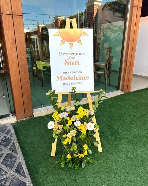 A 'Here comes the Sun' baby shower welcome sign on a wooden easel, decorated with a bouquet of yellow and white flowers.