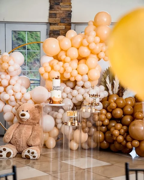 A teddy bear baby shower dessert table with a large brown teddy bear, a neutral balloon arch, and cakes on clear stands.