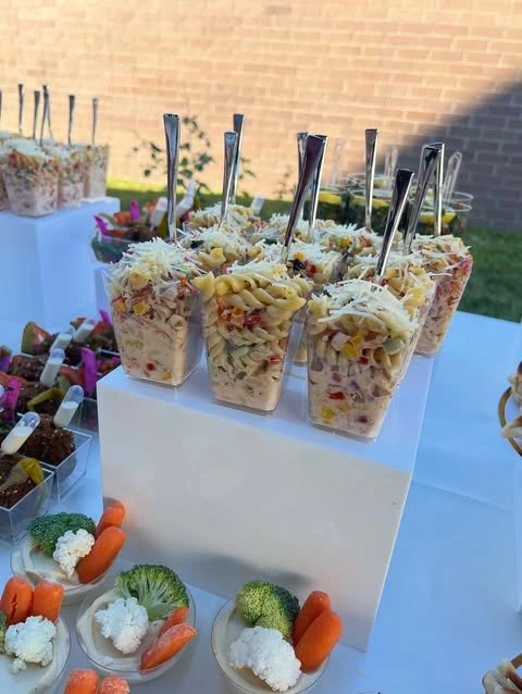 A baby shower food table featuring individual pasta salad cups arranged on a white tiered stand, with veggie cups in the foreground.