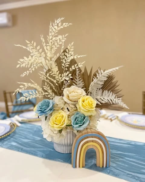 A baby shower centerpiece with yellow and blue flowers, dried ferns, and a small wooden rainbow for a 'Here Comes the Sun' theme.