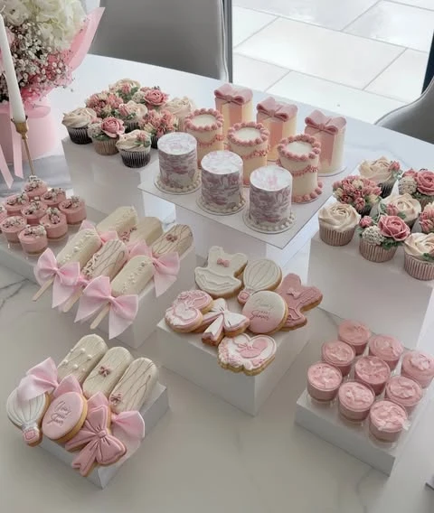 A baby shower dessert table with pink and white treats arranged on clean white multi-level display risers.