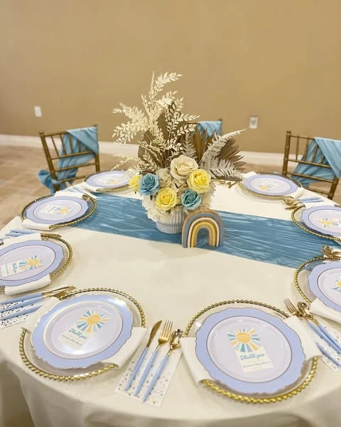 A round table set for a 'Here Comes the Sun' baby shower, showing sun-graphic plates and a floral centerpiece.
