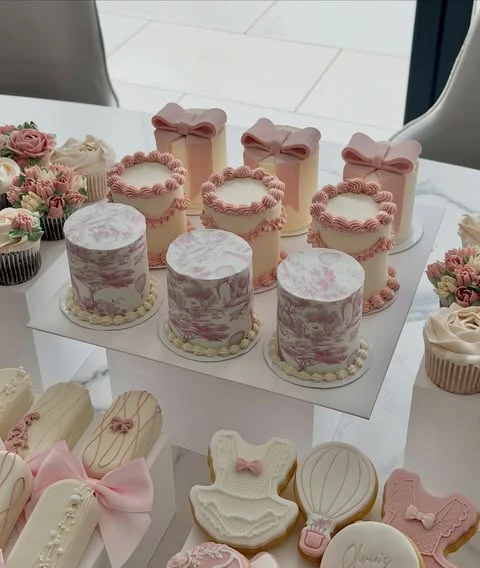 Pink and white mini cakes displayed on tall white risers as part of a baby shower food table setup.