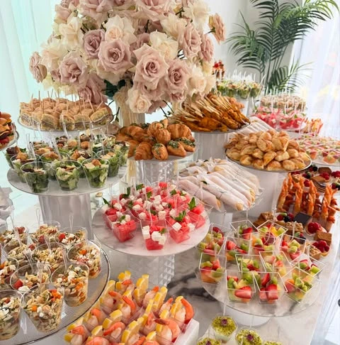 An elegant baby shower food table with tiered platters holding a variety of appetizers in individual serving cups, including salads and shrimp cocktails.