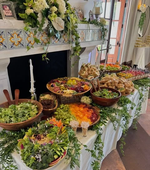 Long baby shower food table with salads, sandwiches, and fruit platters decorated with lush greenery.