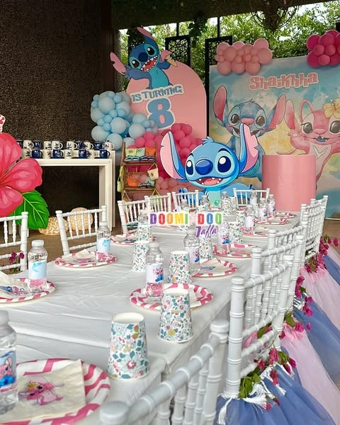 A Stitch and Angel themed birthday party table with a large character backdrop, pink and blue balloons, and themed place settings.