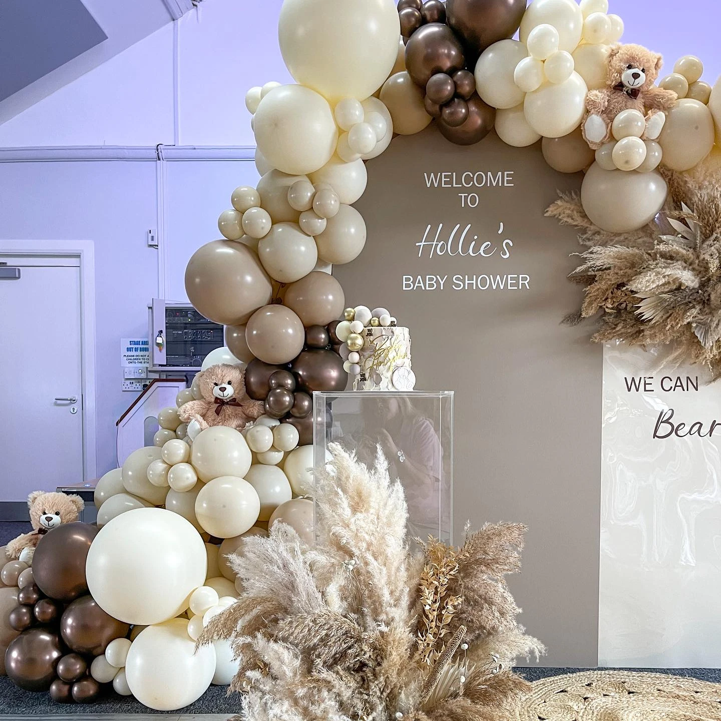 A neutral-toned balloon arch with teddy bears and a 'we can barely wait' sign for a baby shower.