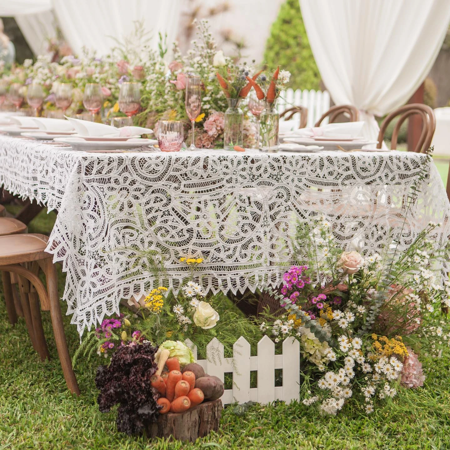 Outdoor baby shower table setting with a white lace tablecloth, wildflower centerpieces, and garden vegetable decor.