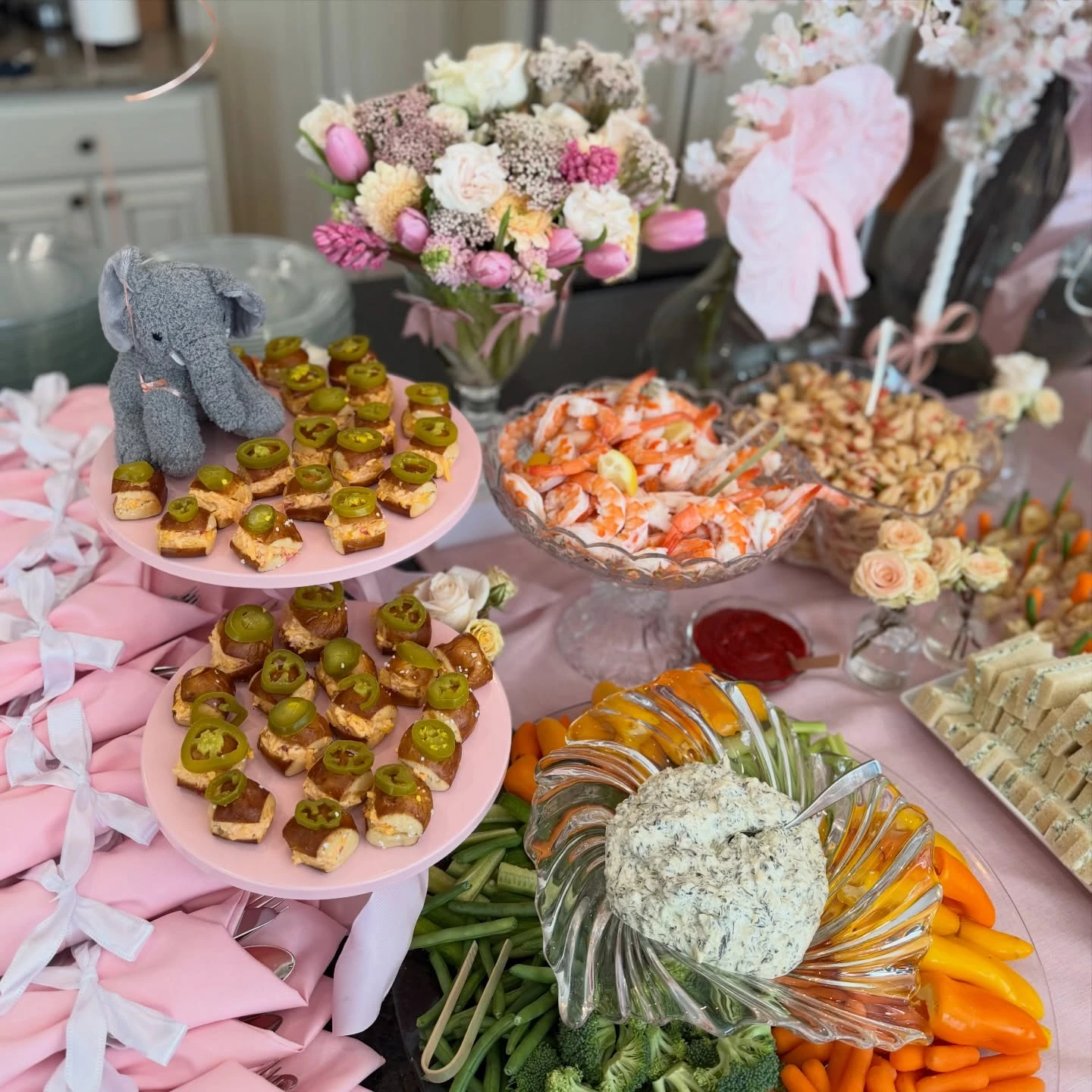 A variety of savory baby shower appetizers including shrimp cocktail, a cheese ball with vegetables, and small bites on a pink stand.