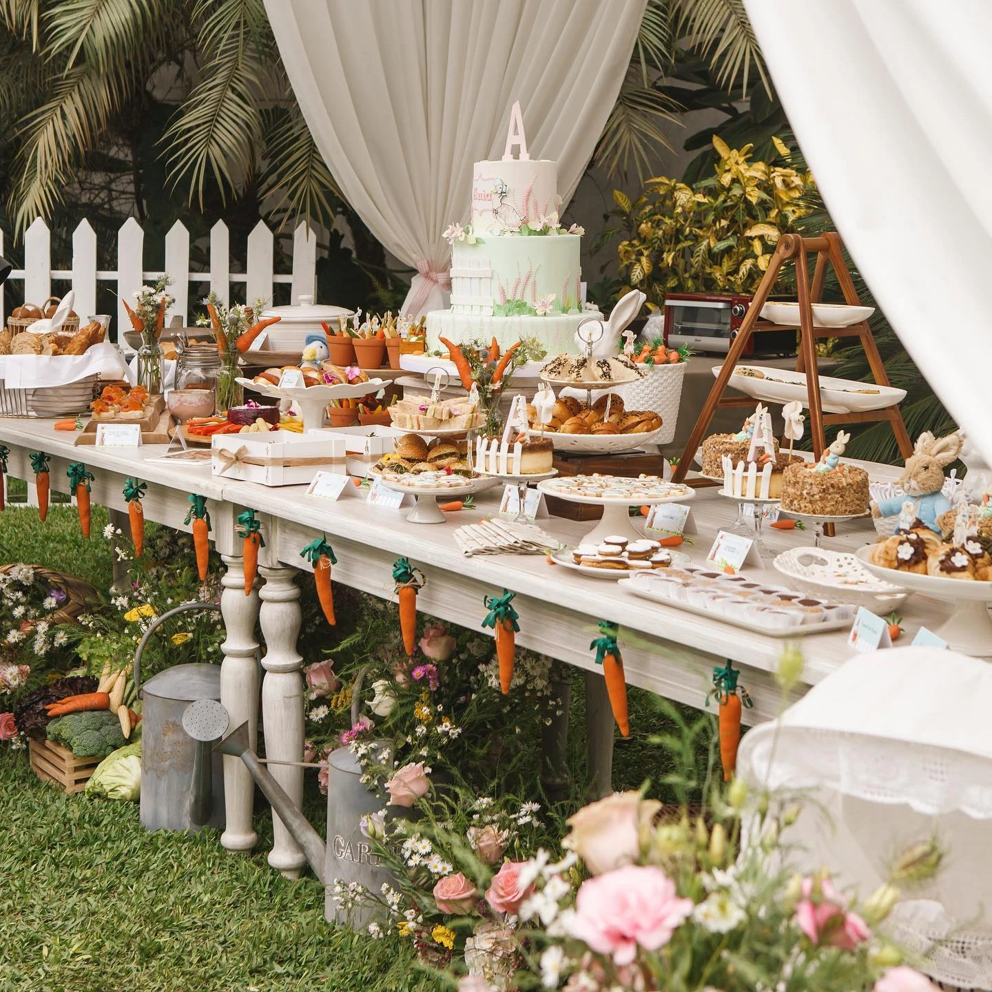 Peter Rabbit baby shower dessert table with a tiered cake, carrot garland, and various pastries in a garden setting.