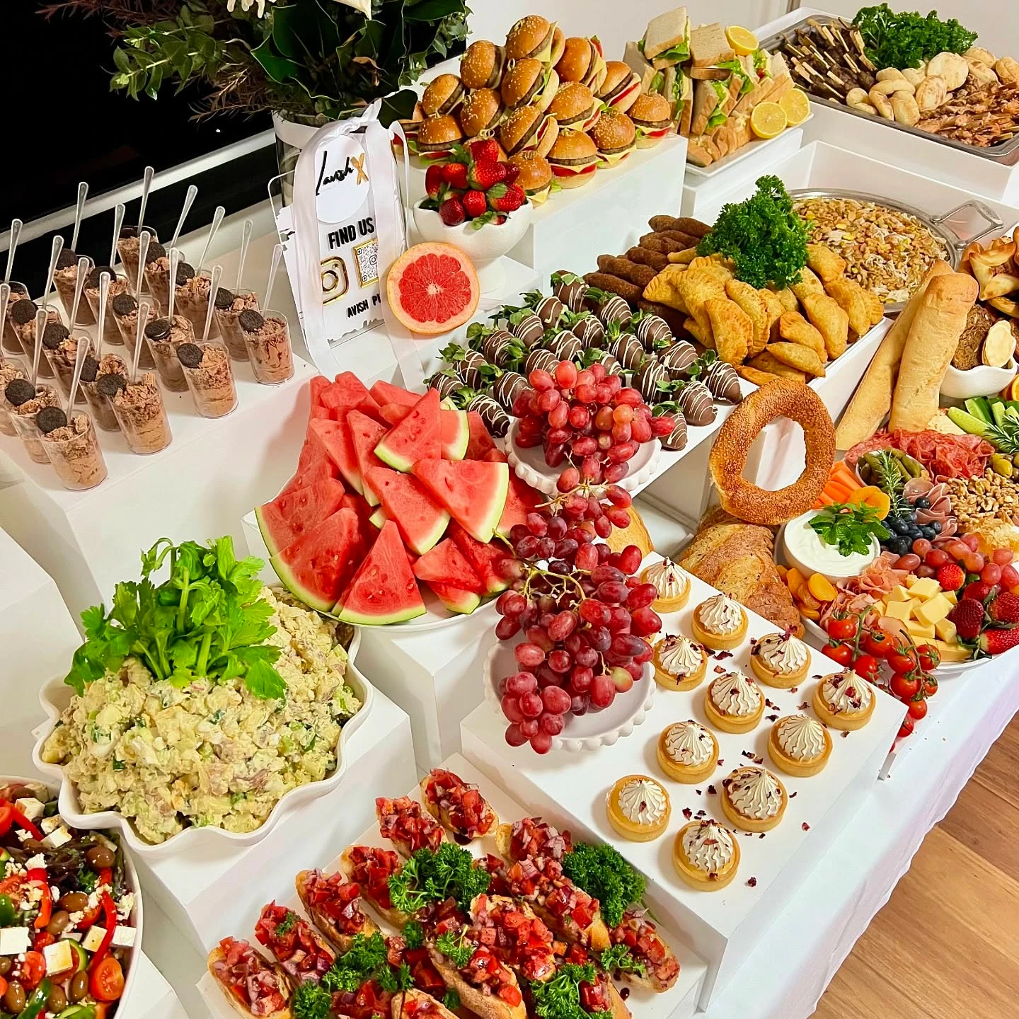A lavish baby shower food table setup featuring a multi-level buffet spread on white risers with a variety of foods like mini sliders, salads, and desserts.