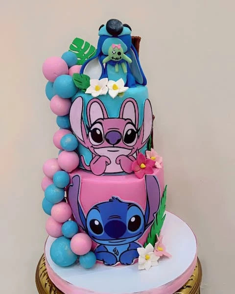 A two-tiered Stitch and Angel themed birthday cake with a pink bottom layer, a blue top layer, and a cascade of pink and blue fondant balls resembling a balloon garland.