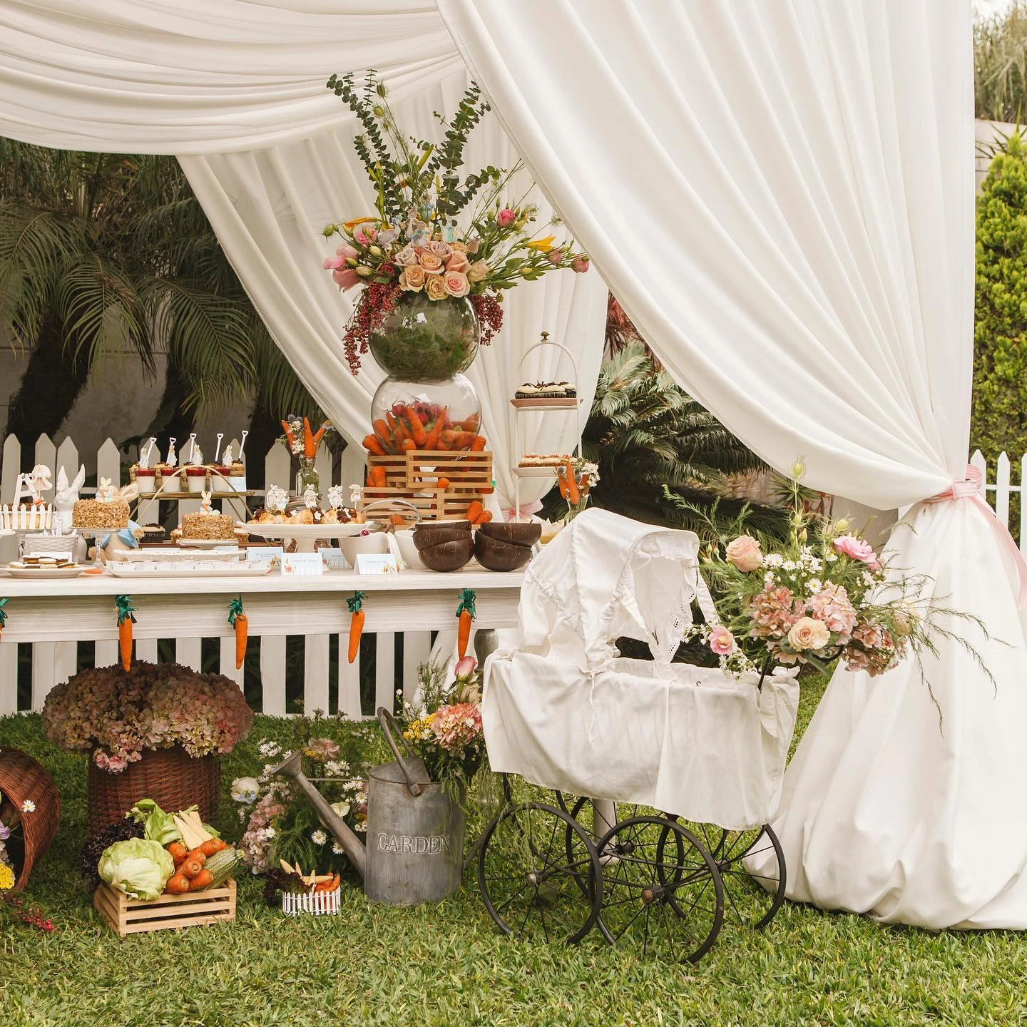 Outdoor baby shower setup with white sheer curtains, a vintage pram, and crates of vegetables.
