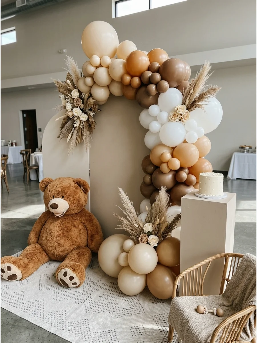 A neutral-toned balloon arch with a large plush teddy bear and dried grass accents.