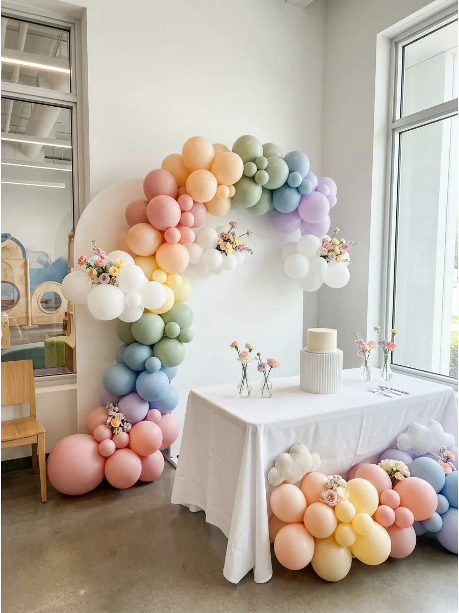 A pastel rainbow balloon garland over a simple white backdrop in a bright room.